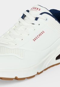 Λευκό sneaker Skechers με κόκκινο λογότυπο, λευκά κορδόνια, εσωτερική επένδυση σε ναυτικό μπλε χρώμα, διακοσμητικές διατρήσεις και σόλα χρώματος καραμέλας.