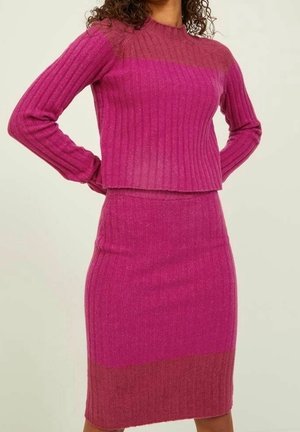 Vrouw draagt een geribbelde, tweekleurige magenta en roze gebreide trui met bijpassende knie-lange rok, staand met handen in de zij.