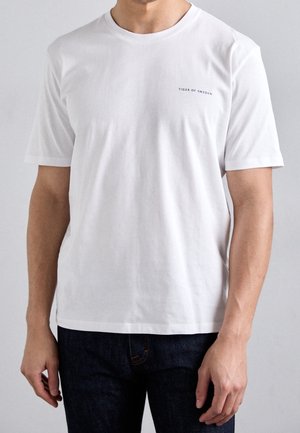 Man draagt een eenvoudige witte T-shirt met korte mouwen en een klein "Tiger of Sweden" logo op de linkerborst, gecombineerd met een donkere spijkerbroek.