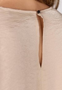 Beige blouse met een textuurafwerking, voorzien van een inkeping aan de achterkant die is vastgezet met een zwarte knop, omgeven door een nette stiklijn.
