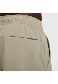 Pantalones cortos beige con cintura elástica, un bolsillo trasero y un pequeño logo "DRI-FIT" bordado encima del bolsillo. Textura de tela suave.