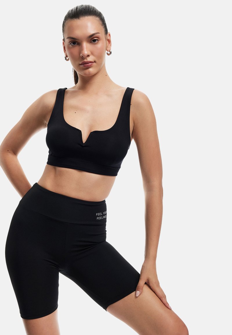 Nainen mustassa V-mallin urheiluliivissä ja korkeavyötäröisissä biker-shorteissa, poseeraa yksi käsi lantion päällä ja toinen reidellä.