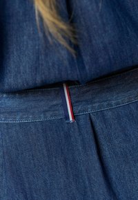 Denimový kúsok s priliehavým pásom a prúžkovaným akcentom v červenej, bielej a modrej farbe. Tkanina má hladkú textúru s viditeľným prešívaním.