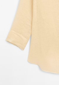 Hellbeige Bluse mit fließendem Design, drei Viertel lange Ärmel, weicher Textur und einem subtilen, leicht unregelmäßigen Webmuster.