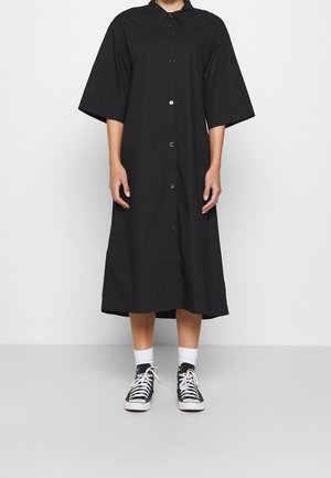 Personne portant une robe noire à boutons mi-longue avec des manches courtes larges et des baskets montantes noires avec des lacets blancs et des chaussettes blanches.