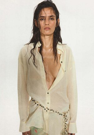 Mujer con el pelo mojado lleva una camisa suelta, parcialmente desabrochada, de color crema y transparente, y un cinturón decorado con lentejuelas circulares brillantes.