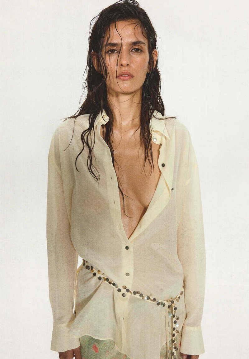 Femme aux cheveux mouillés porte une chemise ample, partiellement déboutonnée, transparente de couleur crème, et une ceinture décorée de sequins ronds brillants.