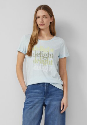 Jeune femme aux longs cheveux portant un t-shirt bleu clair avec le mot « delight » répété en jaune, or et blanc, associé à un jean bleu.