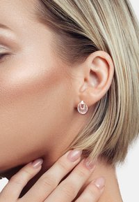 Orecchini a cerchio in oro rosa con un design a conchiglia che presenta linee decorative che si irradiano dal centro. I capelli biondi corti della modella incorniciano il suo viso.