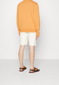 Sudadera naranja, pantalones cortos color crema y sandalias negras con acentos naranjas. Tejido de mezcla de algodón, corte relajado y costuras planas.