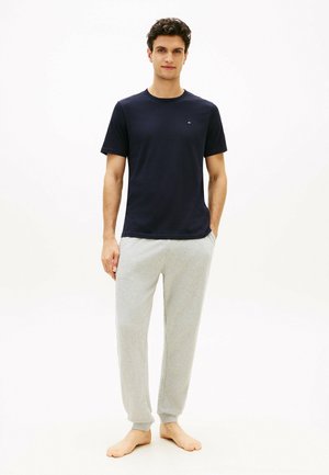 Mand står barfodet iført en marineblå t-shirt med korte ærmer og lysegrå joggingbukser med hænderne i lommerne på en hvid baggrund.