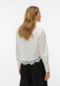 Femme aux longs cheveux blonds portant un chemisier blanc avec une bordure en dentelle florale aux poignets et à l'ourlet, associée à un pantalon noir ample, vue de profil droit.