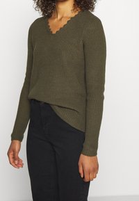 Olivengrüner Strickpullover mit V-Ausschnitt und gewelltem Kragen. Lange Ärmel und leicht asymmetrischer Saum, kombiniert mit schwarzen Jeans.