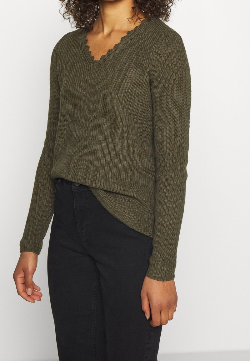 Olivengrüner Strickpullover mit V-Ausschnitt und gewelltem Kragen. Lange Ärmel und leicht asymmetrischer Saum, kombiniert mit schwarzen Jeans.