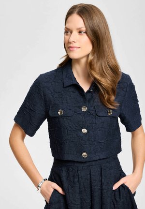 Femme aux longs cheveux châtain clair portant un haut texturé bleu marine à manches courtes boutonné et une jupe taille haute assortie, les mains dans les poches.