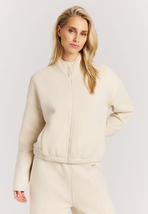 Blonde vrouw die een beige vest met ritssluiting en bijpassende broek draagt, staand met één hand op de heup tegen een effen achtergrond.