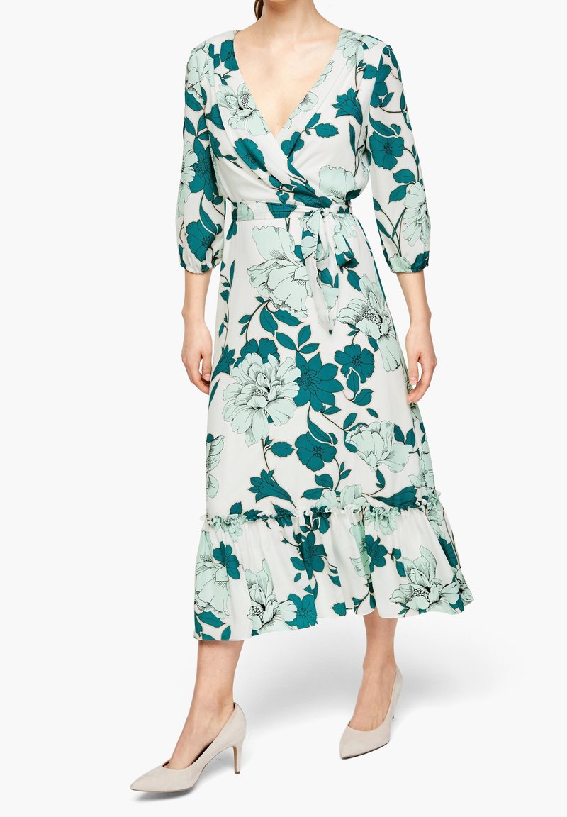 Femme portant une robe portefeuille blanche à imprimé floral turquoise et bleu clair, manches trois quarts, ceinture à la taille, ourlet à volants et talons hauts beiges.