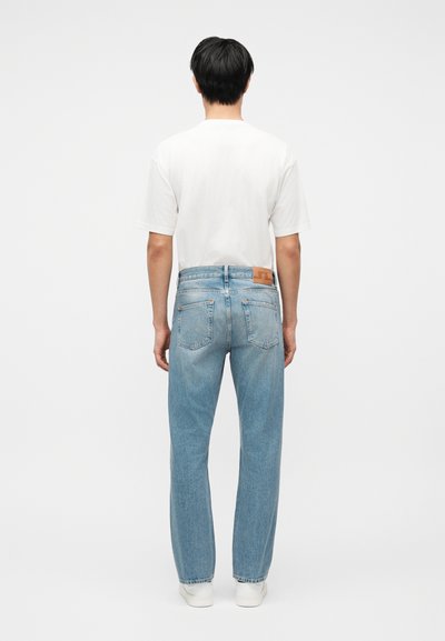 Moški stoji obrnjen stran, oblečen v navadno belo majico, svetlo modre ravne kavbojke in bele superge na belem ozadju.