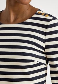 Stripete topp i marineblått og kremhvitt, med rund hals, tre gullknapper på skulderen, og et jevnt, elastisk bomullsmateriale.