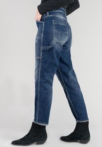 Persona con jeans de mezclilla azules con costura en los bolsillos traseros y botas tobilleras negras, posando frente a un fondo liso.