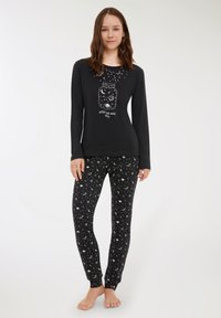 Giovane donna che indossa una maglietta nera a maniche lunghe con un disegno di barattolo e spazio, leggings neri abbinati con un motivo bianco di spazio, in piedi a piedi nudi.