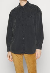 Persona che indossa una camicia nera larga con due taschini sul petto e pantaloni di velluto a coste color senape, in piedi davanti a uno sfondo neutro.