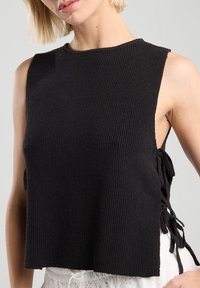 Black cropped tank top with ribbed texture, featuring open sides and tie accents for adjustable fit. Simple, casual design.