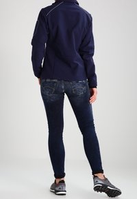 Dunkelblaue Softshell-Jacke mit tailliertem Schnitt und weißen Nähten, kombiniert mit dunkelblauen Jeans und grauen Sneakers, von hinten gezeigt.