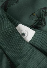 Zaļa T-krekla audums ar ribotu apkakli, kurā ir balta etiķete un caurspīdīga pogas detaļa. Audumam ir teksturēts izskats.