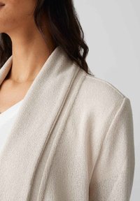 Hellbeiger Cardigan aus strukturiertem Strickstoff, mit einem Schalkragen und lässiger Passform. Sichtbare Steppnähte entlang der Kanten.