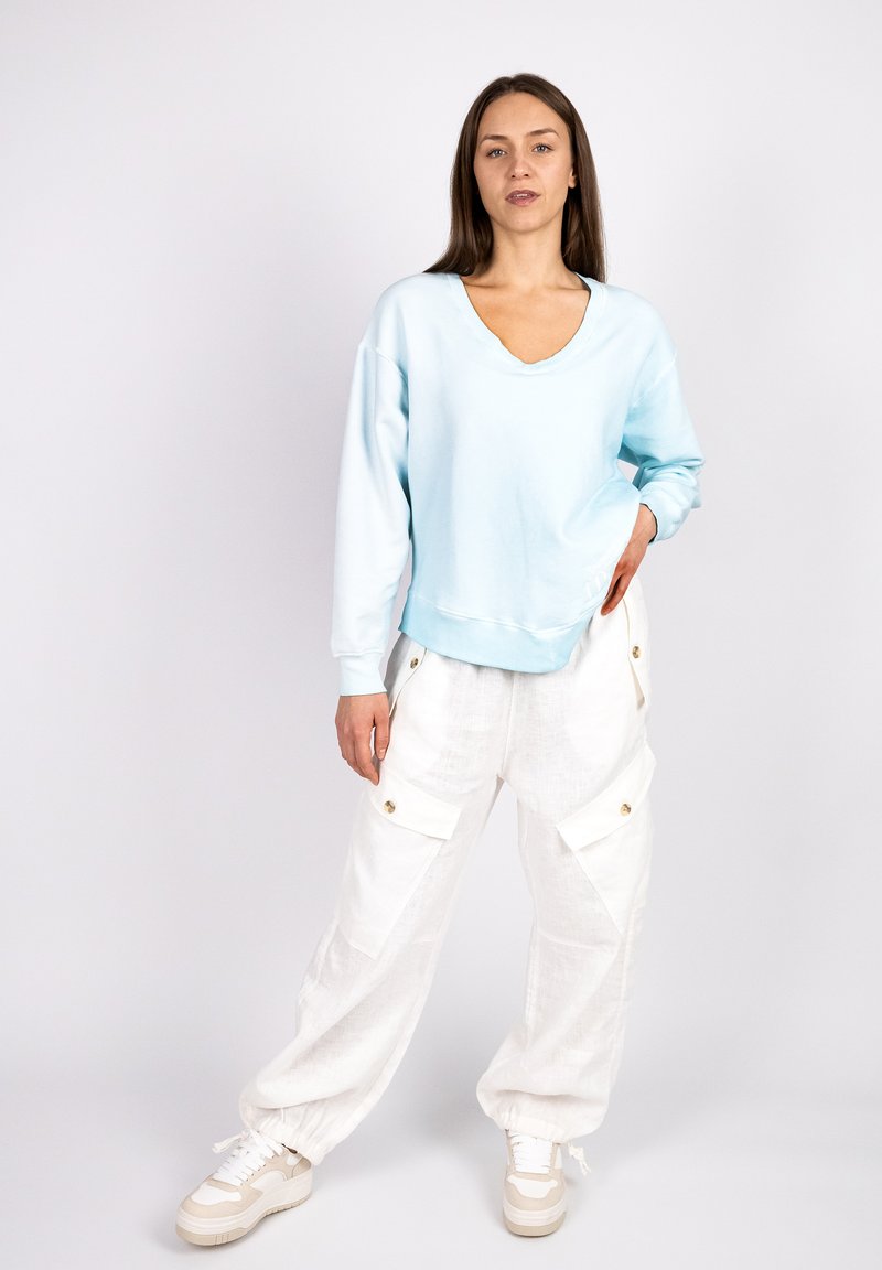 Light blue sweatshirt with a V-neck, oversized fit, and long sleeves. Paired with loose-fitting white cargo pants featuring side pockets and gold buttons.