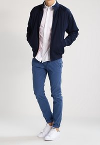 Námořnicky modrá bomber bunda přes bílou košili s puntíkovaným vzorem, doplněná modrými chinos kalhotami a bílými teniskami. Ležérní, vrstvený outfit.