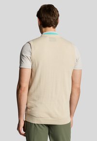 Maglione senza maniche beige con un motivo a maglia testurizzato, caratterizzato da un colletto turchese a contrasto e un orlo a coste, indossato sopra una camicia bianca.
