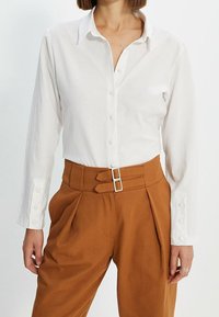 Mujer vistiendo una camisa blanca de botones metida en unos pantalones de color óxido de cintura alta con dobles hebillas.