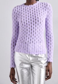 Person som bär en genomskinlig lavendelfärgad långärmad topp med textur, kombinerad med metalliska silverbyxor, sedd från halsen till höfterna.