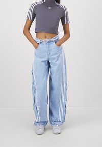 Šedý krátký crop top s krátkým rukávem a vysokým límcem, bílým logem Adidas; světle modré široké cargo džíny s bočními kapsami a bílými pruhy.