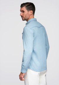 Chemise en denim bleu clair avec manches longues, col boutonné et surpiqûres contrastantes, portée avec un pantalon blanc. Coupe classique et texture lisse.