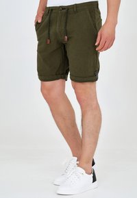 Shorts vert olive en tissu texturé, fermeture à boutons et cordons noirs. Ourlet retroussé et poches latérales. Portés avec des baskets blanches.