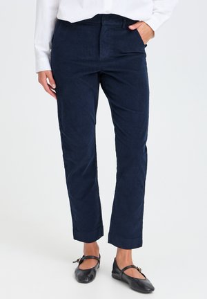 Personne portant un pantalon droit bleu foncé, une chemise blanche et des ballerines Mary Jane noires avec une bride et une boucle. Une main dans la poche.