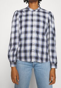 Camicia a quadri con bottoni in nero, bianco e blu, con maniche a sbuffo, colletto alto e orlo dritto con chiusura a bottone.