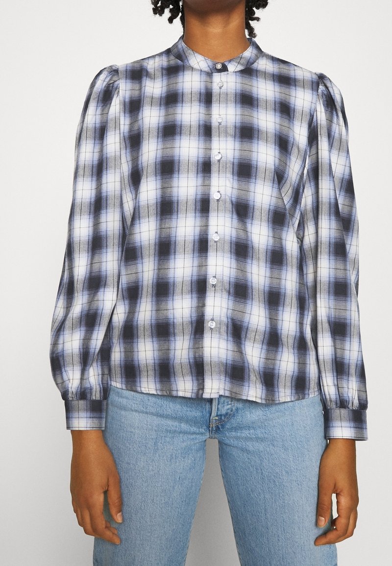 Camicia a quadri con bottoni in nero, bianco e blu, con maniche a sbuffo, colletto alto e orlo dritto con chiusura a bottone.