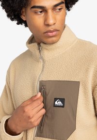 Tan fleecejacka med hög krage, dragkedja och en brun bröstficka med en logotyp patch. Texturerat, varmt tyg.