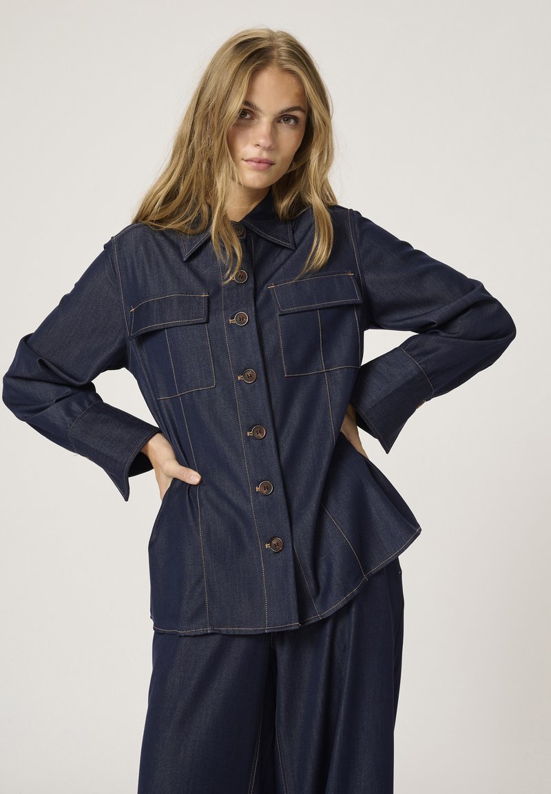 Cămașă din denim de culoare albastră închisă, cu guler ascuțit, două buzunare pe piept, nasturi maro și cusături în contrast. Prezintă o tiv flared și mâneci structurate.