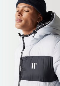 Chaqueta acolchada en gris claro con un panel negro que presenta el logo "11°". Capucha ajustable con cordones y tirador de cremallera de marca.