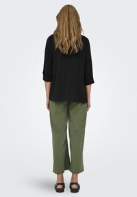 Camicia nera testurizzata con maniche arrotolate, abbinata a pantaloni larghi verde oliva. Il modello indossa sandali neri, visto da dietro.