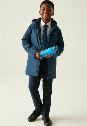 Młody chłopiec uśmiechający się, ubrany w niebieski płaszcz, białą koszulę, paskowany krawat, ciemne spodnie i czarne buty, trzymający niebieską butelkę na wodę.