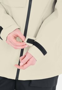 Beige wasserdichte Jacke mit schwarzem Klettverschluss am Ärmel und Reißverschluss. Nahaufnahme von Händen, die den Ärmel anpassen; glatte Textur und funktionales Design sichtbar.