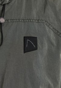 Lekka oliwkowa kurtka z teksturowanej tkaniny, z kapturem na sznurku oraz czarną naszywką z białym haftem na klatce piersiowej.