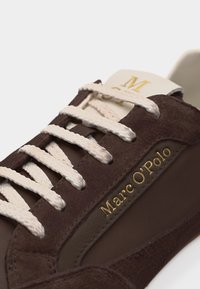 Καφέ σουέτ αθλητικό παπούτσι με λευκά κορδόνια και χρυσά γράμματα «Marc O'Polo» στη πλευρά, εμφανιζόμενο σε κοντινή γωνία.