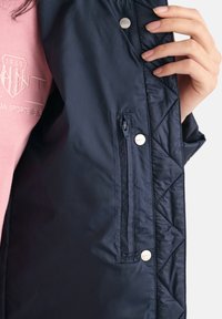 Hand hält eine offene marineblaue Jacke, die eine mit Reißverschluss gesicherte Innentasche und Druckknöpfe zeigt. Darunter wird ein rosa Hemd mit besticktem Text getragen.